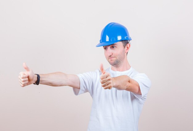 Tシャツ、ヘルメットで二重の親指を見せて、陽気に見える建設労働者