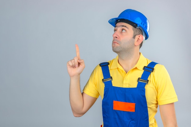 Construction worker in uniform and blue safety helmet surprised looking up and pointing up with finger isolated on white