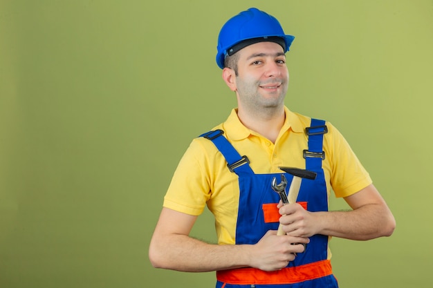 Free photo construction worker in uniform and blue safety helmet with smile on face standing with hammer isolated on green