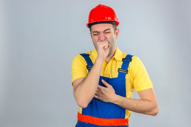 Construction worker in uniform and red safety helmet bored yawning tired coning mouth with hand isolated on white