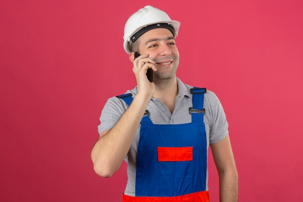 Muratore in uniforme e casco di sicurezza con il sorriso sul fronte che parla dal telefono cellulare isolato sul rosa