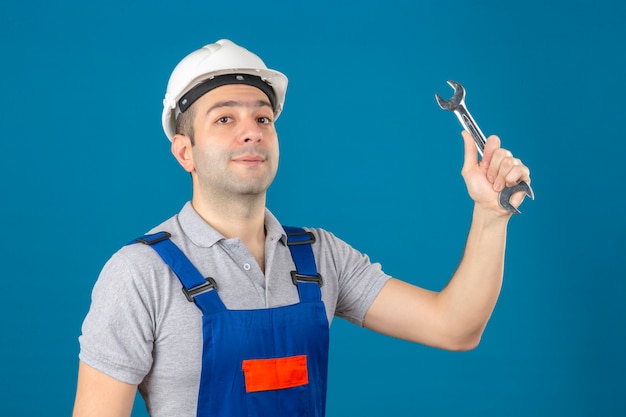 Construction worker in uniform and safety helmet with wrench in raising hand on blue isolated