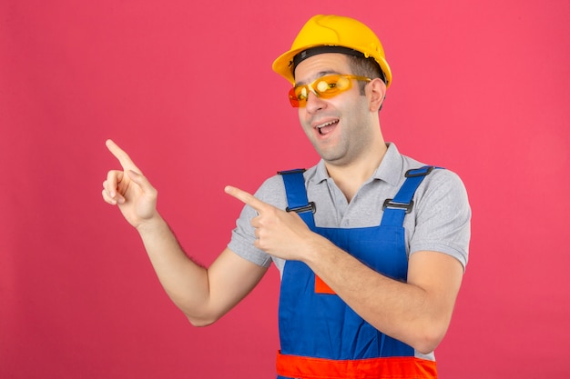 制服の安全ヘルメットとメガネを着て笑顔とピンクに分離された側に2本の手と指で指している建設労働者