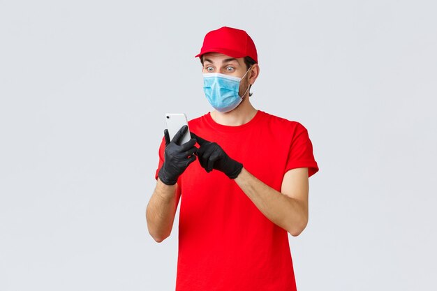 Contactless delivery, payment and online shopping during covid-19, self-quarantine. Excited courier in face mask and gloves, staring astonished at smartphone display, reading order info