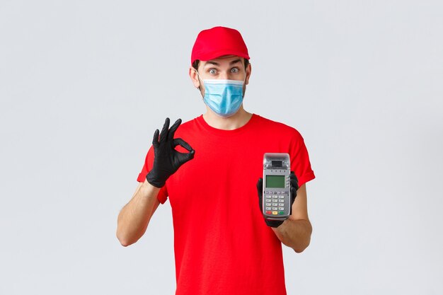 Contactless delivery, payment and online shopping during covid-19, self-quarantine. Excited courier in red uniform, face mask and gloves recommend pay order with POS terminal and credit card