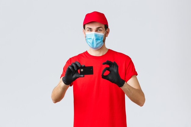 Contactless delivery, payment and online shopping during covid-19, self-quarantine. Friendly courier in red uniform, cap and t-shirt, wear face mask and gloves, thumb-up recommend credit card paying