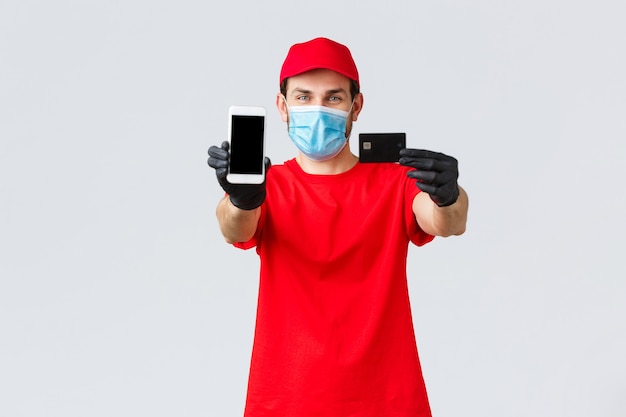 Contactless delivery, payment and online shopping during covid-19, self-quarantine. Friendly courier in red uniform, face mask and gloves, showing smartphone screen and credit card, order internet.