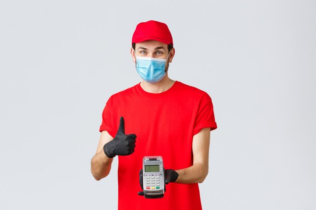 Contactless delivery, payment and online shopping during covid-19, self-quarantine. Friendly smiling courier in red uniform cap, t-shrit, medical mask and gloves, advice pay order with POS terminal