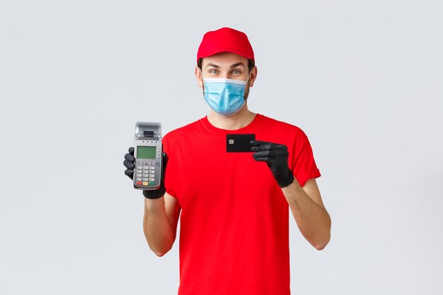 Contactless delivery, payment and online shopping during covid-19, self-quarantine. Handsome courier with paying terminal POS and credit card, provide safe pay order, wear face mask and gloves
