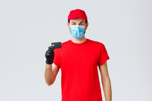 Contactless delivery, payment and online shopping during covid-19, self-quarantine. Young courier in red uniform cap, face mask and gloves, showing credit card, easy paying and ordering