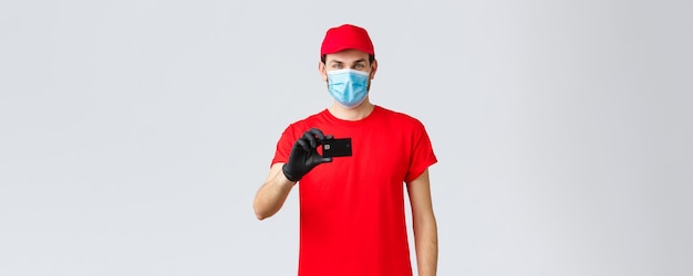 Contactless delivery payment and online shopping during covid19 selfquarantine Handsome courier in red uniform cap medical face mask and gloves show credit card order internet