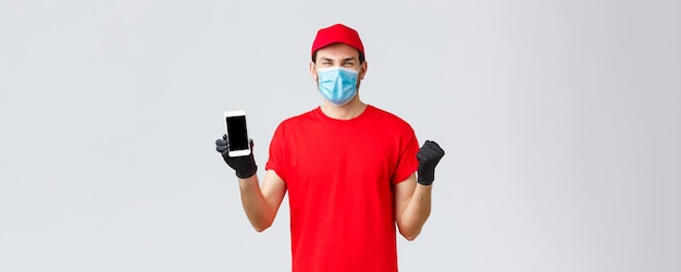 covid19自己検疫中の非接触型決済とオンラインショッピング素晴らしいプロモーションショーのスマートフォンのスクリーンウェアマスクを祝う赤いユニフォームキャップTシャツで陽気な宅配便を喜ぶ