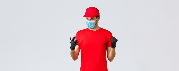 Free photo contactless delivery payment and online shopping during covid19 selfquarantine enthusiastic courier in red uniform face mask and gloves rejoicing over good news look smartphone screen