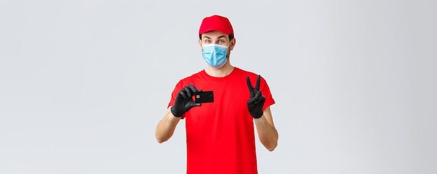 Free photo contactless delivery payment and online shopping during covid19 selfquarantine friendly courier in red uniform gloves and face mask provide safe paying option show credit card and peace sign