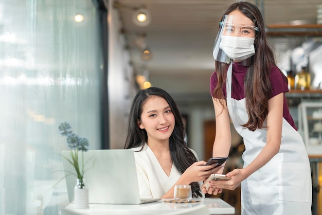 Contactless payment with mobile in restaurant Waitress standing wearing protective face mask and apron with asian smiling customer with her smartphone on hand about to pay by qr code payment system