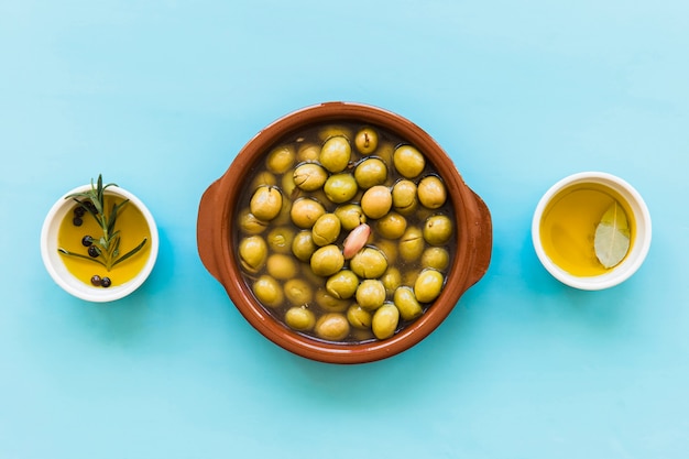 Container of green olives with bowl of oils on blue background