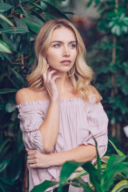 Contemplated blonde young woman standing near the green plants