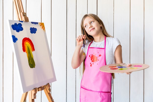 Contemplated girl holding palette and paintbrush in hand standing near the easel