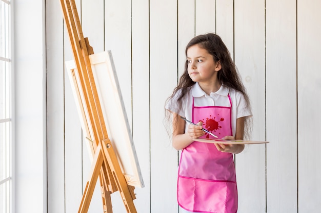 Free photo contemplated little girl with pink apron holding paintbrush and palette