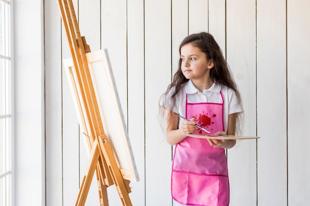 Contemplated little girl with pink apron holding paintbrush and palette