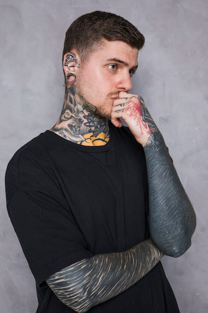 Contemplated tattooed young man with piercing in his ear standing against grey wall