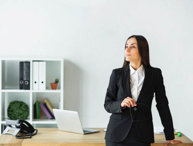 Contemplated young businesswoman holding eyeglasses in hand standing behind the office desk