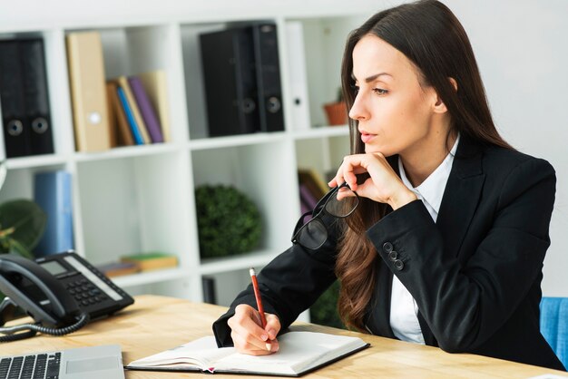 Contemplated young businesswoman holding pencil over diary on the office desk