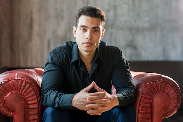 Free photo contemplated young man sitting on red armchair with hand clasped against wall