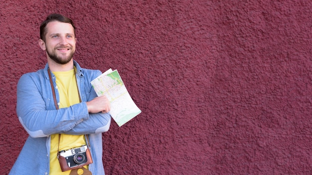 Contemplating young smiling traveler photographer holding map standing near textured wall background