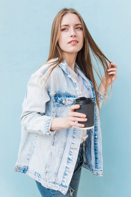 Созерцая молодая женщина, держащая одноразовые кофе, стоя перед синей стеной