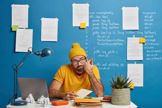 Contemplative bearded student makes notes in notepad during remote work with laptop computer, eats delicious sandwich, writes down ideas for creating own web site