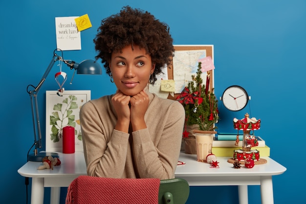 Free photo contemplative curly woman keeps hands under chin, looks thoughtfully aside, poses against workplace decorated for christmas