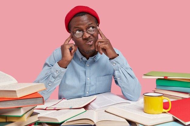 Contemplative dark skinned college student keeps both fingers on temples, purses lips and looks with hesitaton aside, tries analyze information in his mind, being workaholic