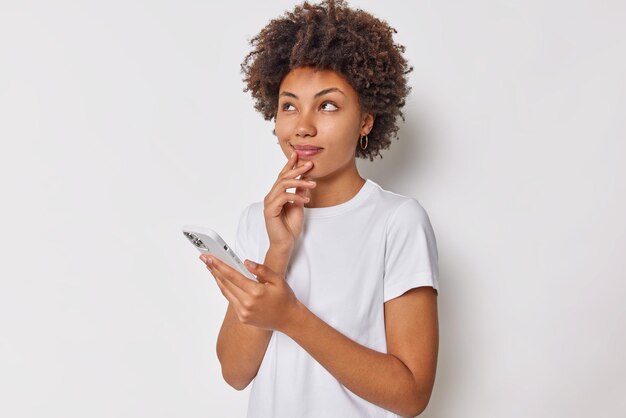 Contemplative dreamy woman looks with thoughtful expression above thinks what answer to give on received message holds mobile phone uses application wears t shirt isolated over white background