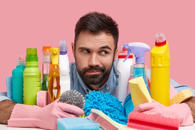 Contemplative man with dark stubble, wears rubber gloves, poses near many detergents, holds sponge, going to wash dish, scrubs bathtub