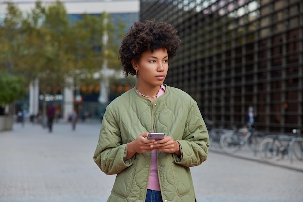 La giovane donna contemplativa con i capelli ricci tiene le moderne reti di cellulari social media in città indossa pose giacca