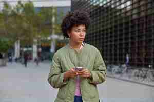 Foto gratuita la giovane donna contemplativa con i capelli ricci tiene le moderne reti di cellulari social media in città indossa pose giacca