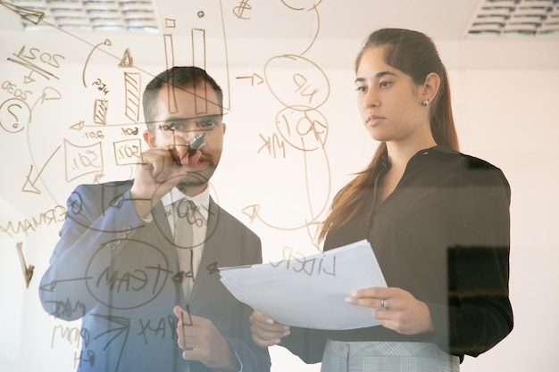 Content African American manager writing chart on glass board. Professional young pretty female colleague holding document and looking at graph in conference room. Teamwork and marketing concept