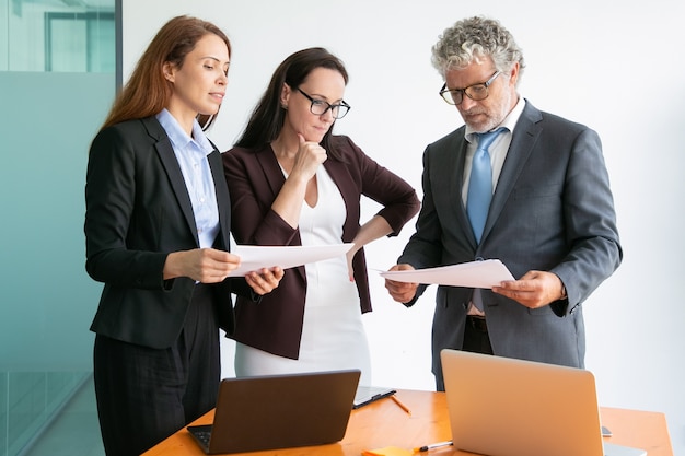 Content businesspeople discussing project, watching documents and standing together near table with laptops