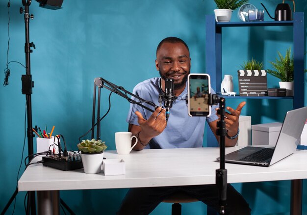 Content creator interacting with fanbase in studio looking at live video podcast setup sitting at desk. Smiling vlogger talking with audience in front of recording smartphone during online live show.