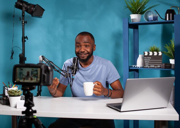 Content creator talking to followers in online morning show in front of live vlog setup with filming video camera on tripod. Smiling vlogger interacting with audience sitting at desk in studio.