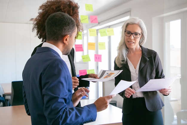 Content female manager explaining statistics data to colleagues. Professional business partners listening experienced grey-haired businesswoman in conference room. Teamwork and management concept