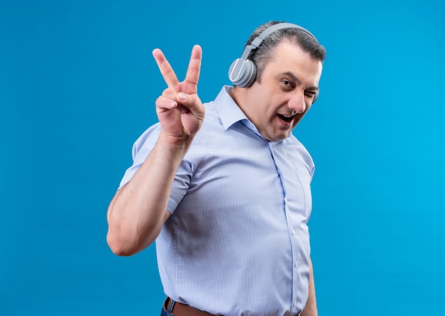 Content middle-aged man in blue striped shirt in headphones looking into the camera and hand gesture with raised up two fingers on a blue space