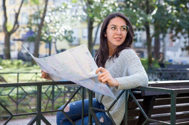 Content pretty young woman using paper map on bench outdoors