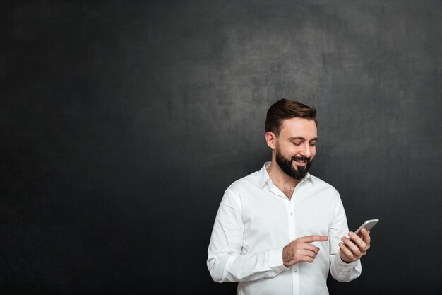 テキストメッセージを入力するか、濃い灰色の上のスマートフォンを使用してソーシャルネットワークでフィードをスクロールする白いシャツのコンテンツの笑みを浮かべて男