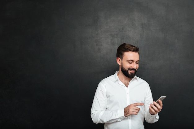 Content smiling man in white shirt typing text message or scrolling feed in social network using smartphone over dark gray
