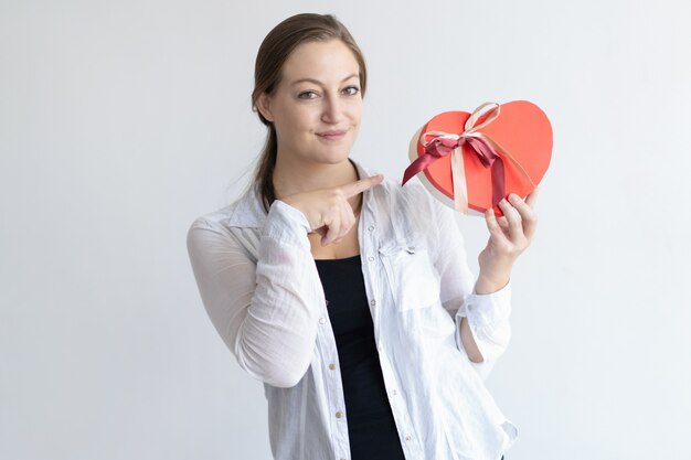 Content woman holding heart shaped gift box and pointing at it