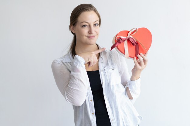 Содержимая женщина держа коробку подарка в форме сердца и указывая на ее