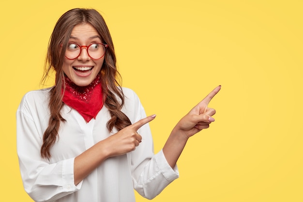  content woman with European appearance, has joyful expression, indicates with both index fingers, shows free space for your advertising content. Joyful girl in white shirt and bandana