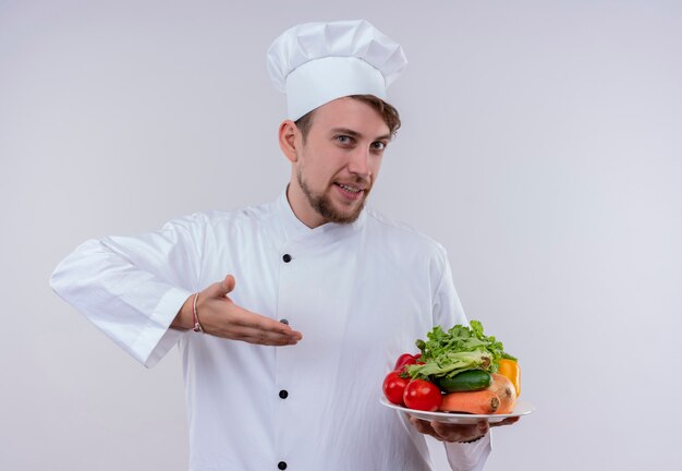 흰 벽에 토마토, 오이, 양상추와 같은 신선한 야채와 함께 하얀 접시를 들고 흰 밥솥 유니폼과 모자를 쓰고 콘텐츠 젊은 수염 난 요리사 남자